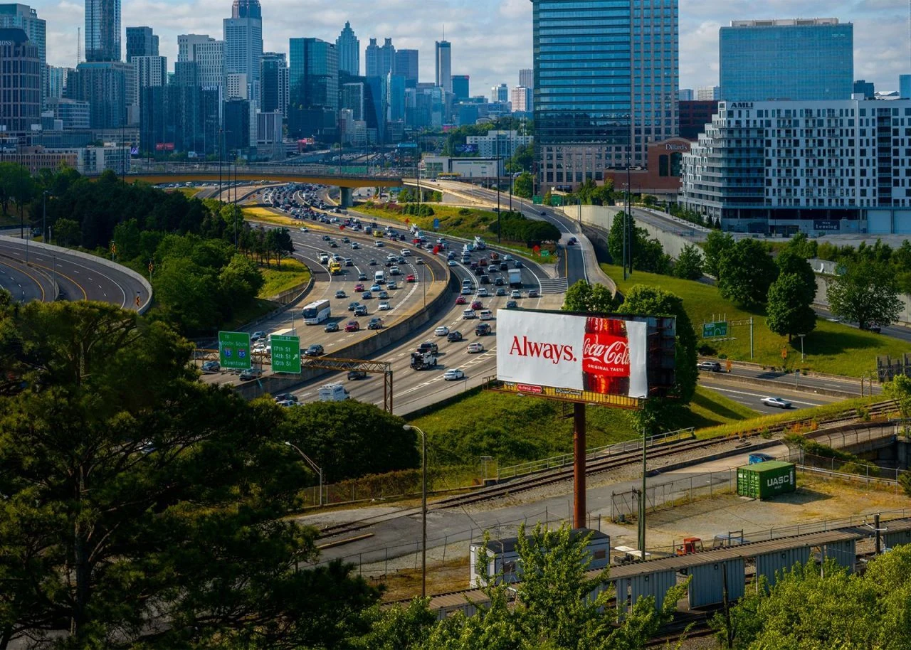 Coca-Cola roadside billboard using the word Always beside a close-up of a Coca-Cola bottle overlooking a busy urban highway.
