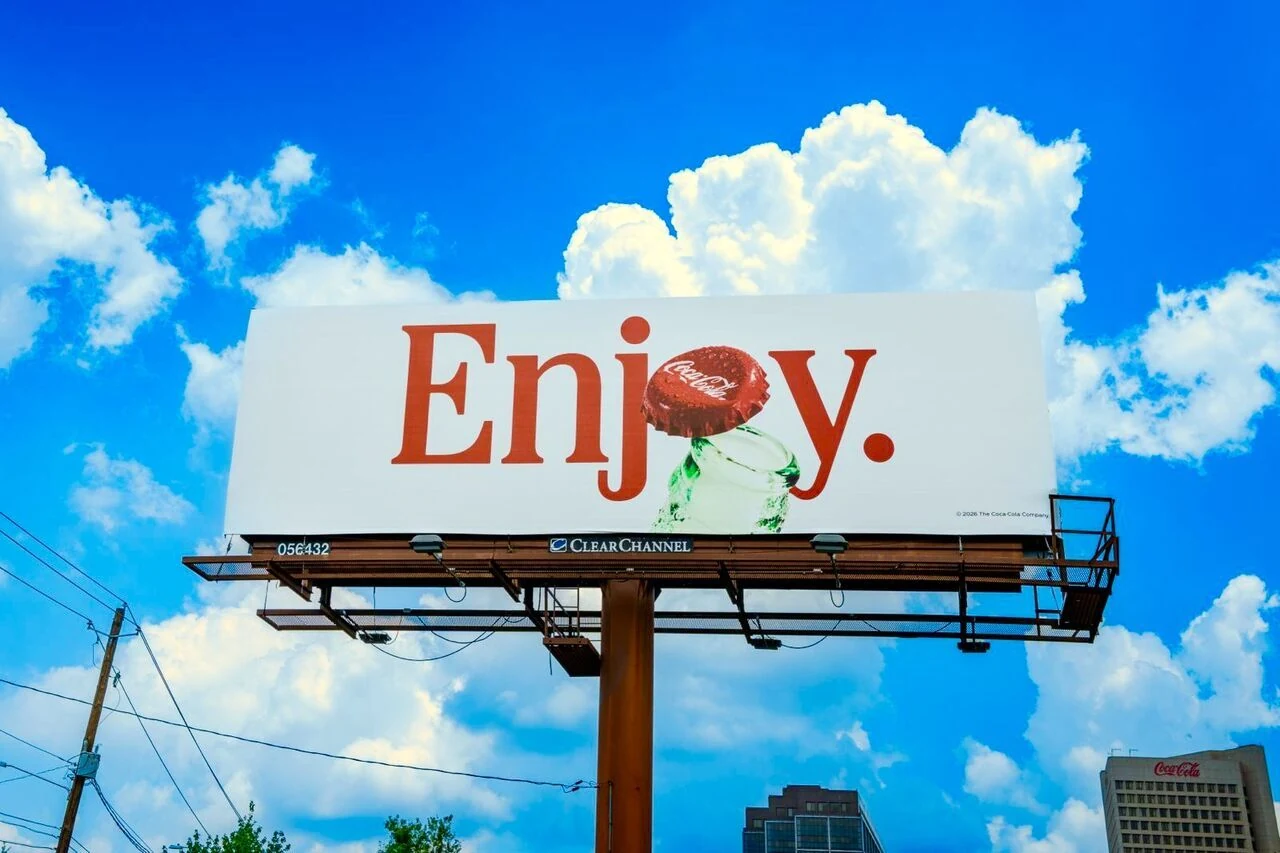 Minimal Coca-Cola billboard showing the word Enjoy with a Coca-Cola bottle cap lifting from a glass bottle against a white background.