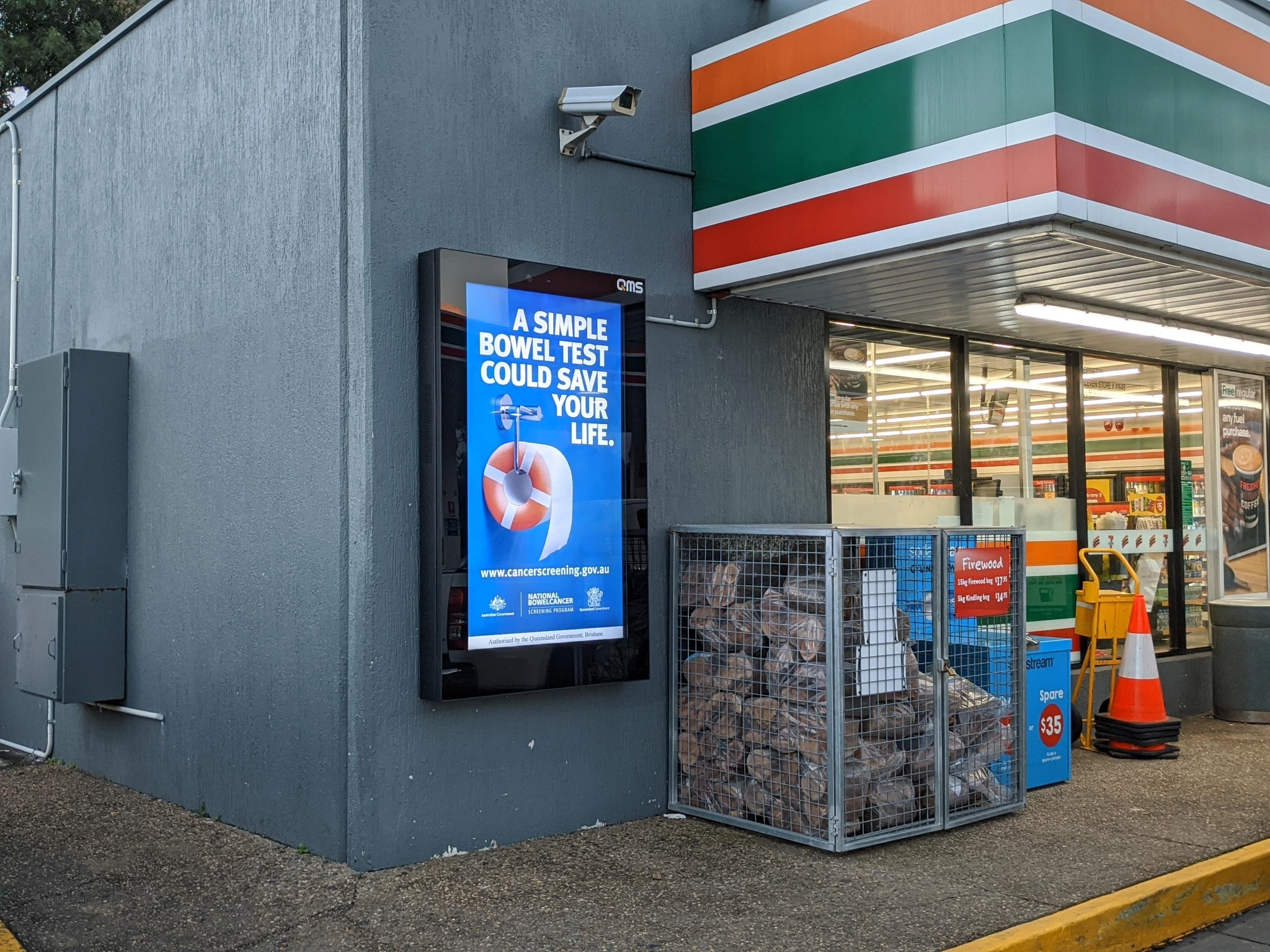 Digital out-of-home screen outside a convenience store and gas station showing a bowel screening public health message.