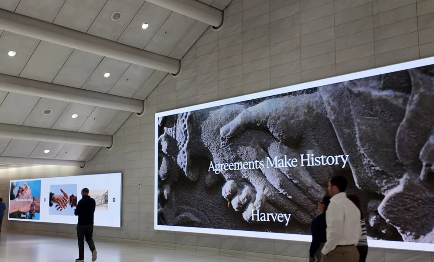 Harvey Agreements Make History campaign displayed on large digital screens in a modern transit or terminal concourse.