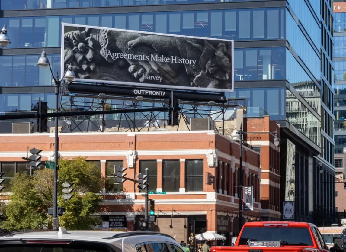 Harvey Agreements Make History black-and-white billboard displayed above a busy city street.