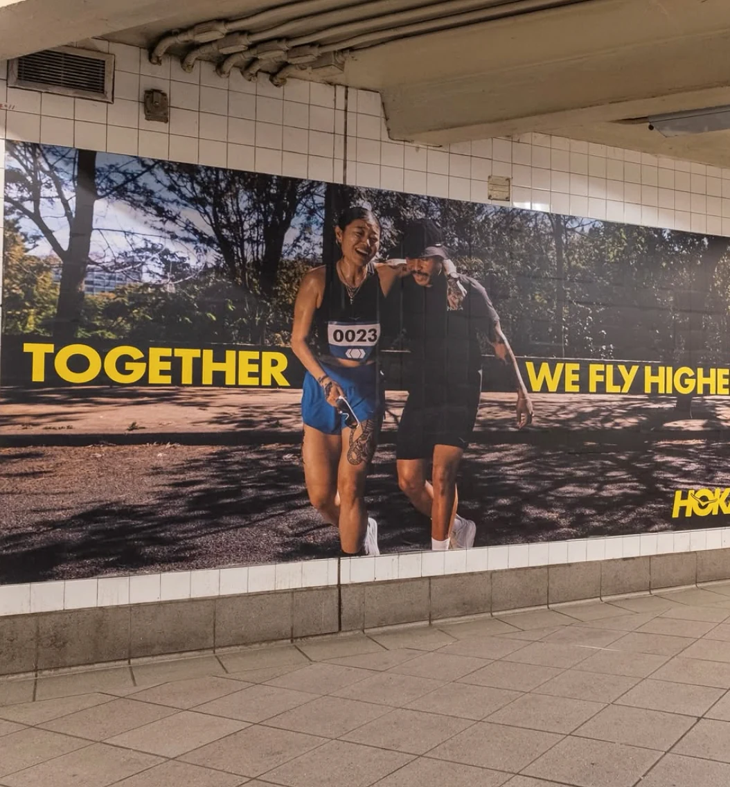 HOKA Together We Fly Higher large wall advertisement inside the New York City subway featuring two runners supporting each other.