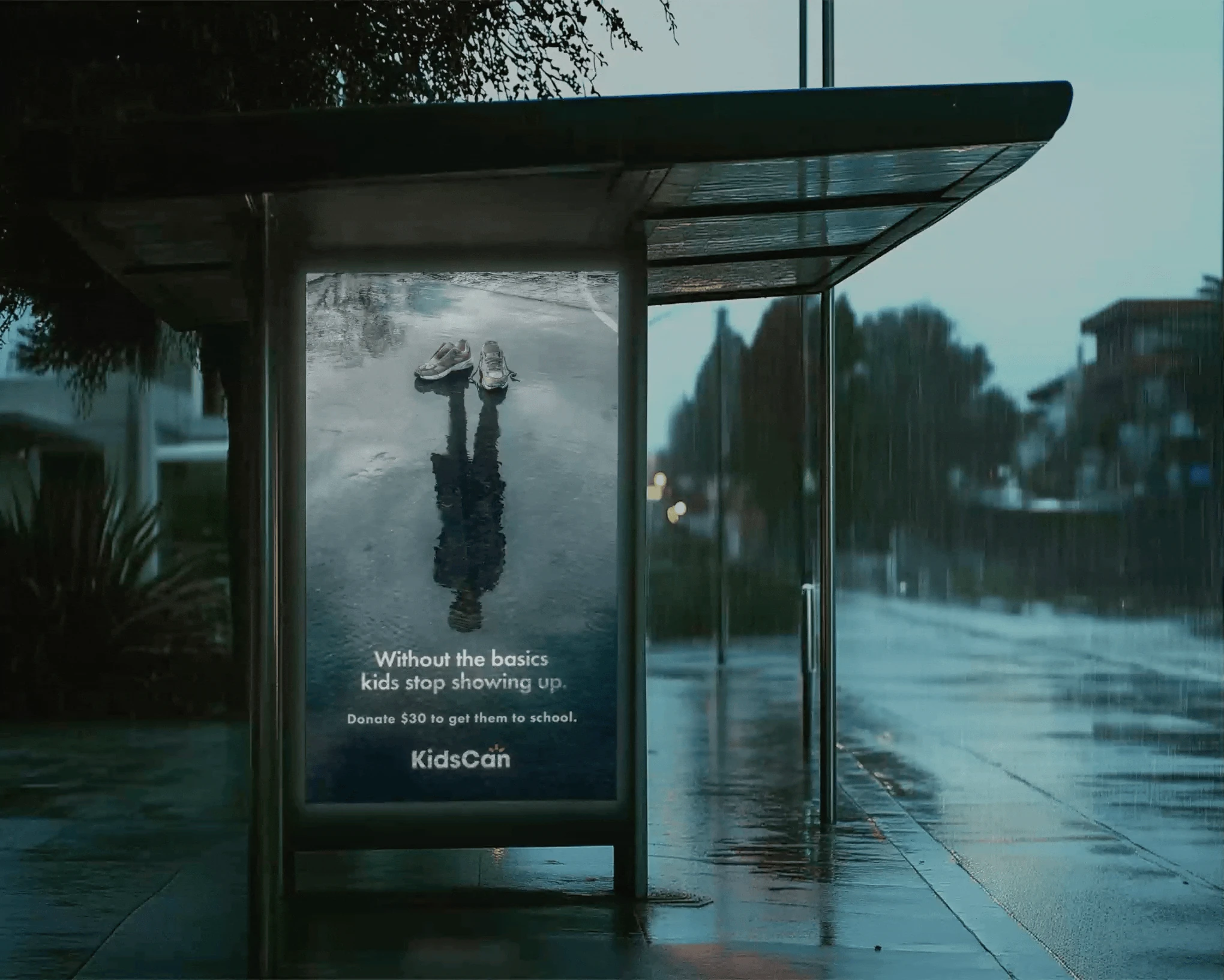KidsCan Reflections bus shelter poster displayed in rainy weather, showing children’s shoes and a reflected silhouette with a call to donate for school essentials.
