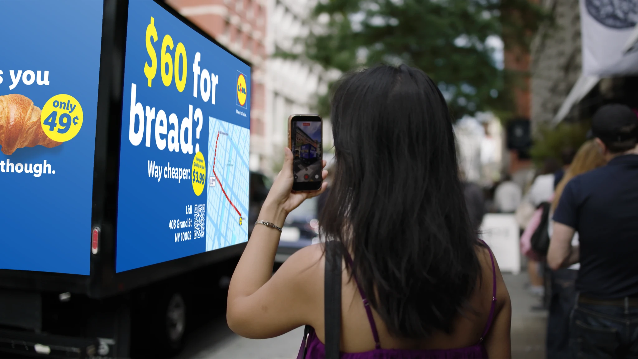 Passerby photographing a Lidl digital billboard truck in Manhattan showing that 60 dollars can buy 122 croissants at Lidl.