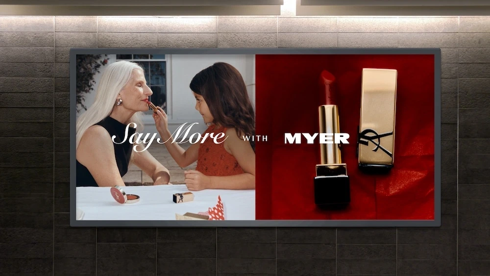 Myer Christmas billboard showing a granddaughter applying lipstick to her grandmother beside premium beauty product imagery.