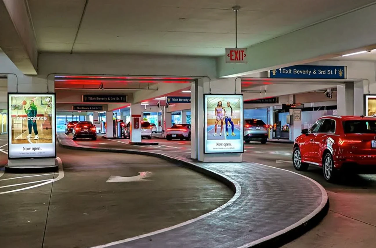 Multiple illuminated New Balance advertisements positioned throughout The Grove parking structure beside arriving vehicles.