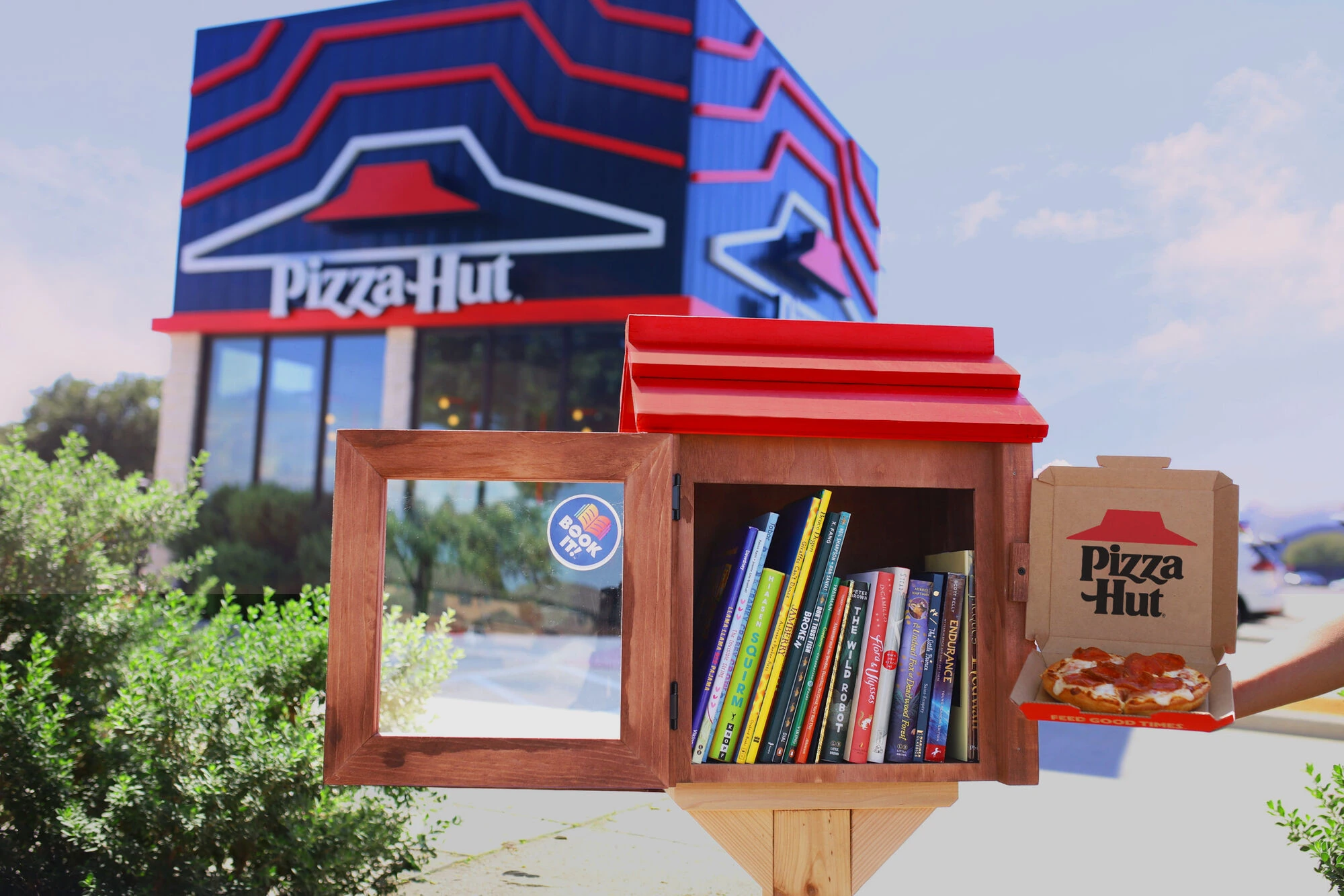 Pizza Hut Little Hut Library with its red roof open and stocked with books beside a Pizza Hut pizza box in front of a Pizza Hut restaurant.