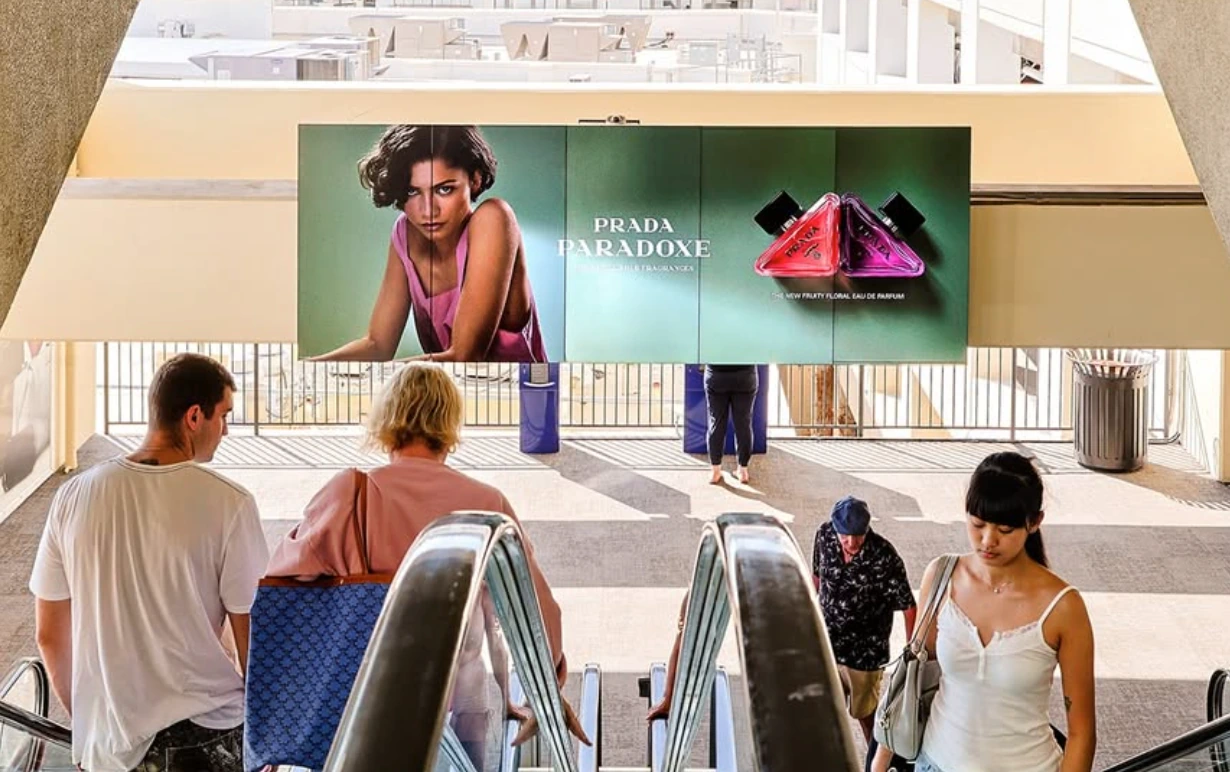 Wide Prada Paradoxe digital spectacular above an escalator at The Grove featuring the campaign portrait and triangular perfume bottles.