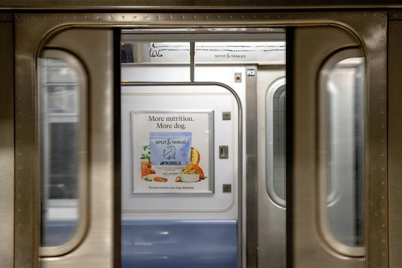 Spot and Tango UnKibble subway poster visible through the open doors of a New York City train car.