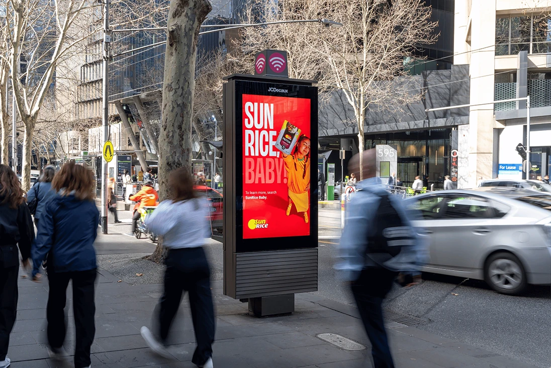 SunRice Protein Rice Baby digital outdoor advertising in Australia promoting Microwave Protein+ with a bold red creative execution in a busy urban environment.